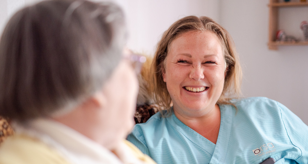 Närbild på  en äldre kvinna som och pratar och skrattar med en kvinna från personalen på äldreboendet. 