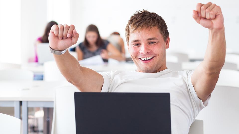 En ung glad kille sitter framför sin dator, andra ungdomar sitter i bakgrunden.