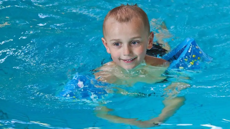 En liten pojke simmar med armkuddar i en inomhusbassäng.