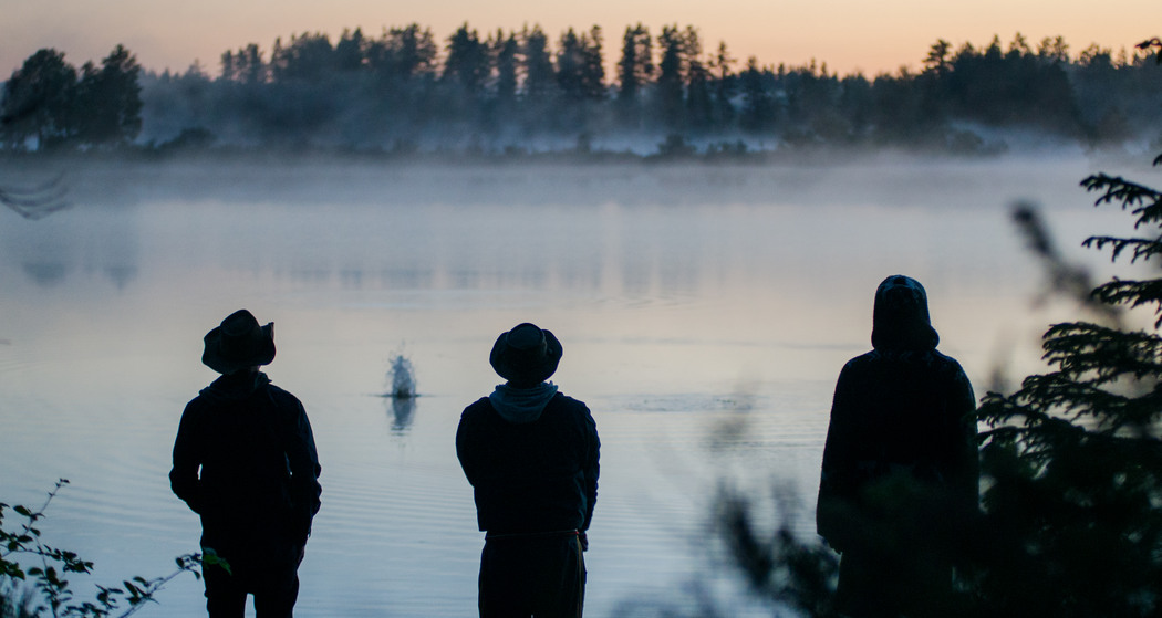 Tre personer står vid en sjö och kastar sten i motljus så det är endast deras siluetter som syns..