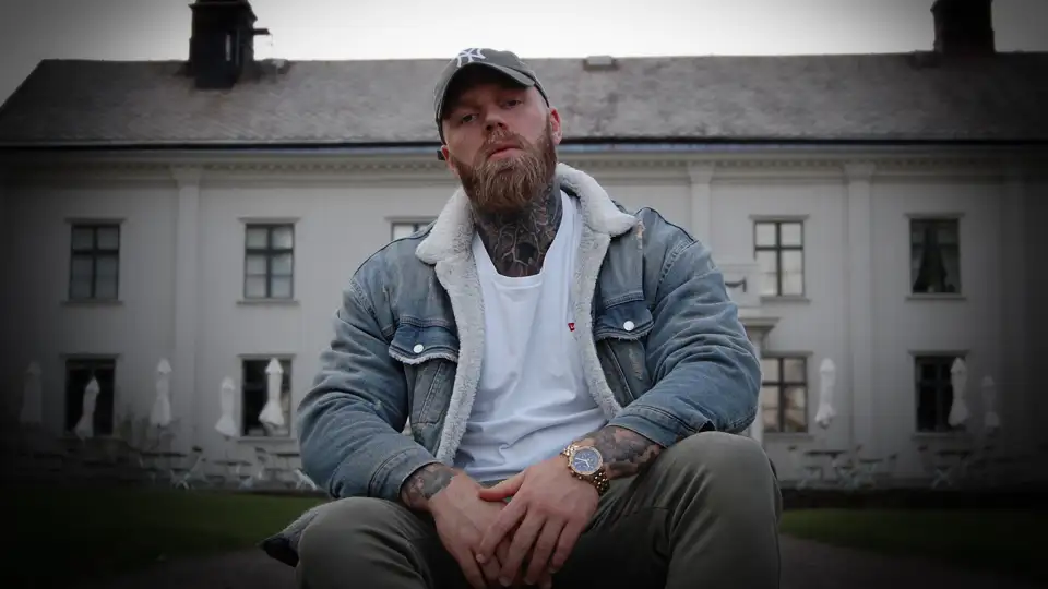 En man med skägg som sitter framför en byggnad.