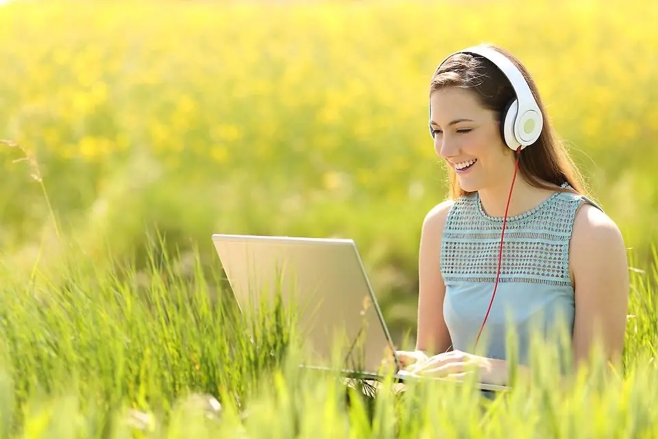 Ung tjej sitter i en grönskande åker med en laptop och hörlurar,