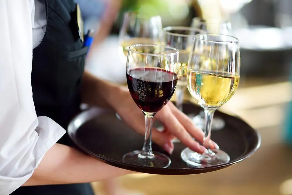 Servitris bär på en rund bricka med fem vinglas, fyra fyllda med vitt vin och ett med rött vin. Foto.