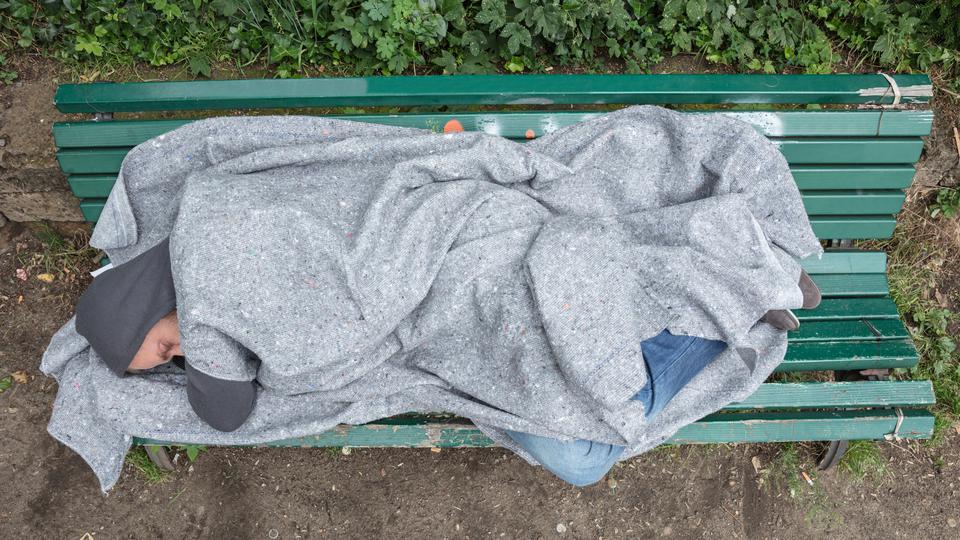 Hemlös man ligger på en grön parkbänk och sover. Foto.