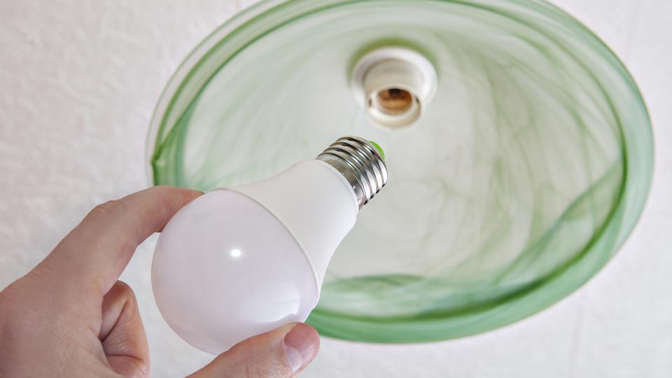 Hand håller i en LED-lampa som ska bytas i en grön taklampa. Foto.