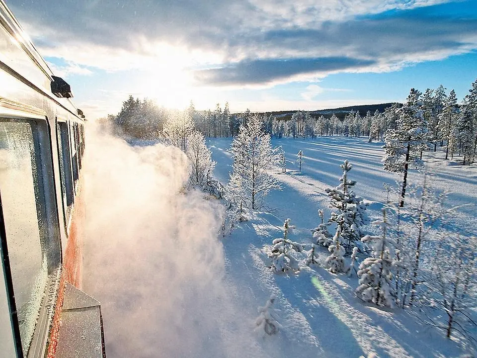 Inlandsbanetåget åker i ett vinterlandskap med snörök och solsken.