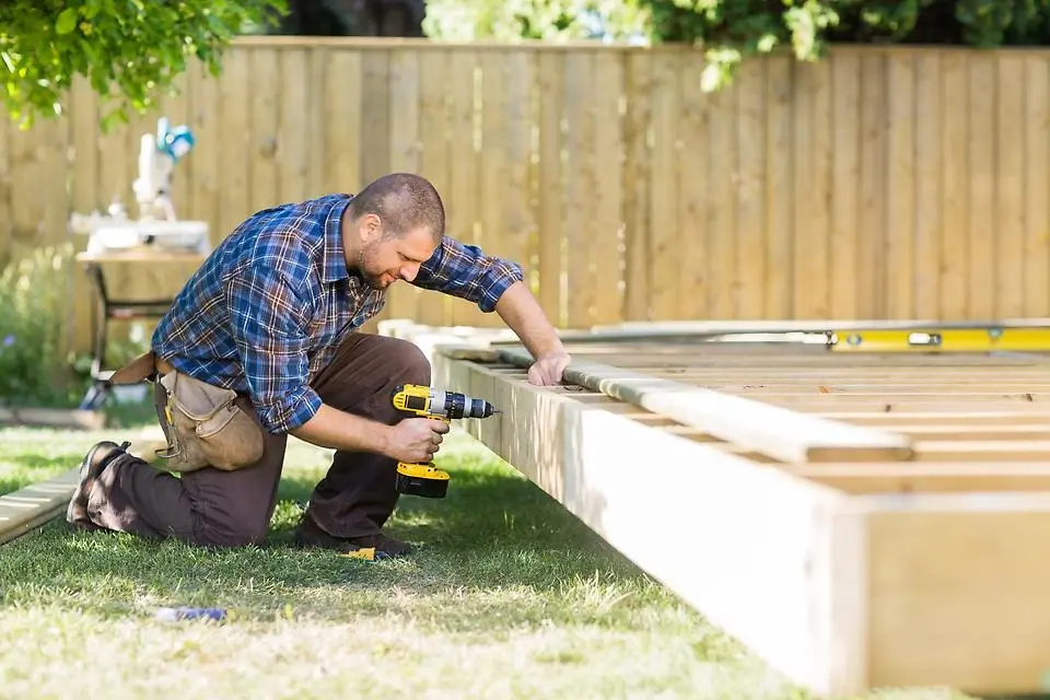 En man bygger grunden till en byggnad. Han står på knä på gräset och borrar i en träbjälke. Foto.