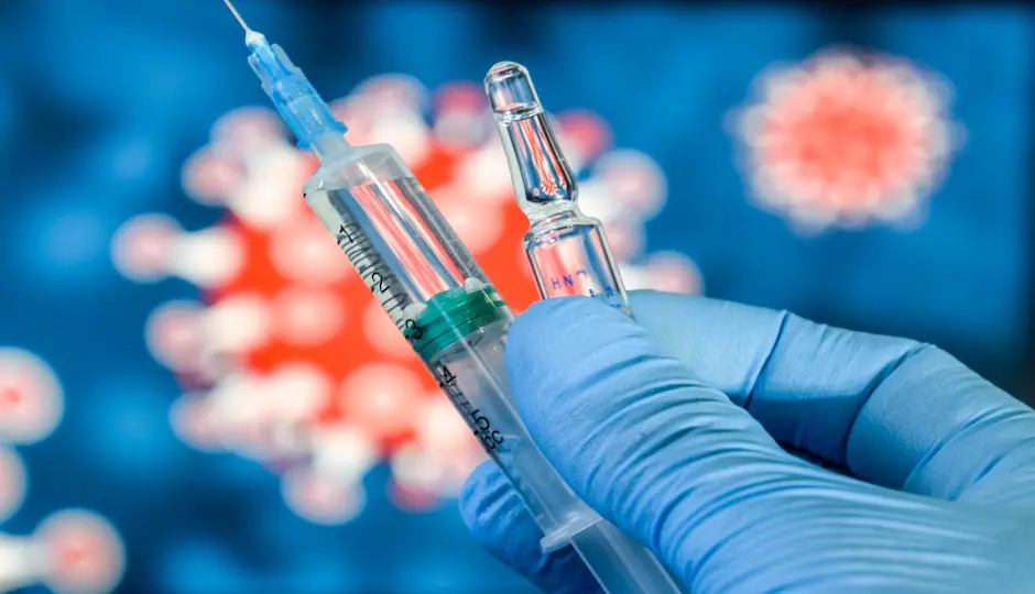 En hand med blå handske håller i vaccinspruta. I bakgrunden bild på coronavirus i microskåp. Foto. 