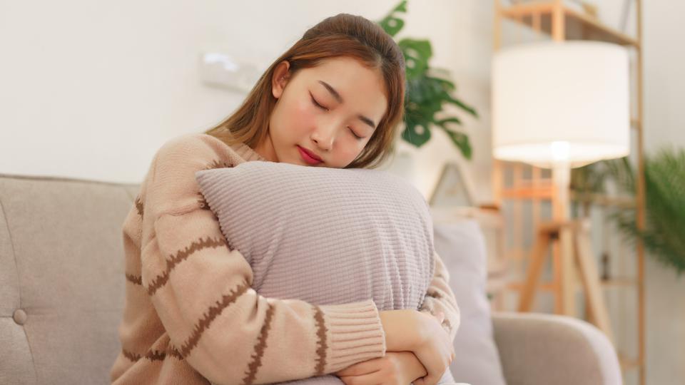 En ung kvinna sitter blundande  i en soffa och kramar en kudde. Foto.