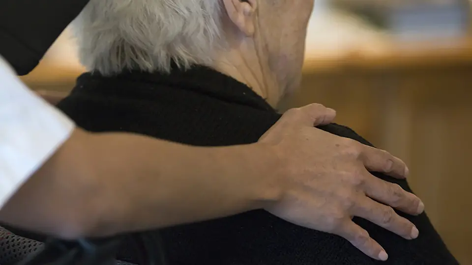 Äldre kvinna i rullstol, personal lägger sin hand på kvinnans rygg.