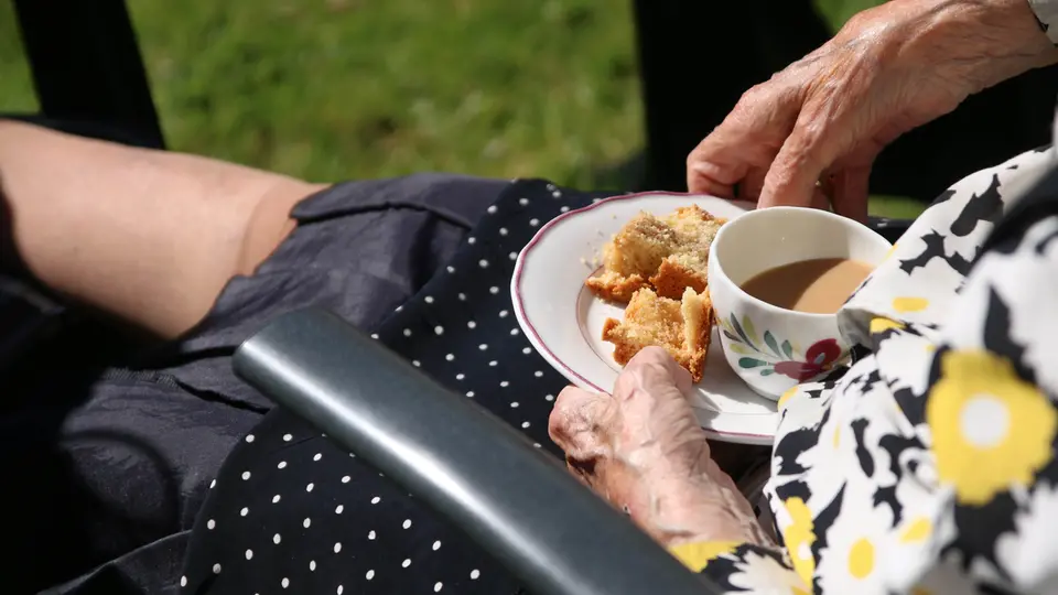 Äldre person dricker kaffe ute i solen. Händer som håller fat med kaka och en kaffekopp. 