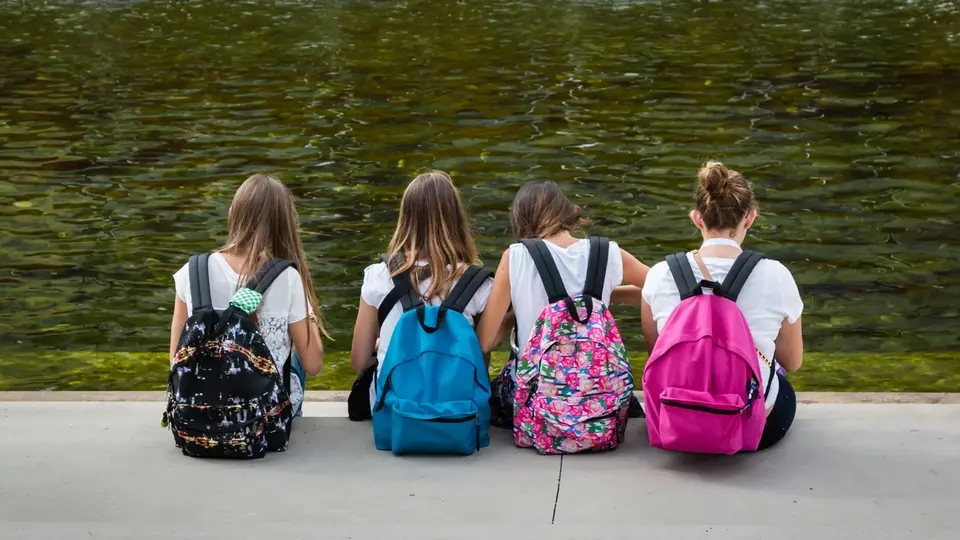 Elever på utflykt. De sitter i en trapp vid en å. Alla har färgglada ryggsäckar på sig. Foto.