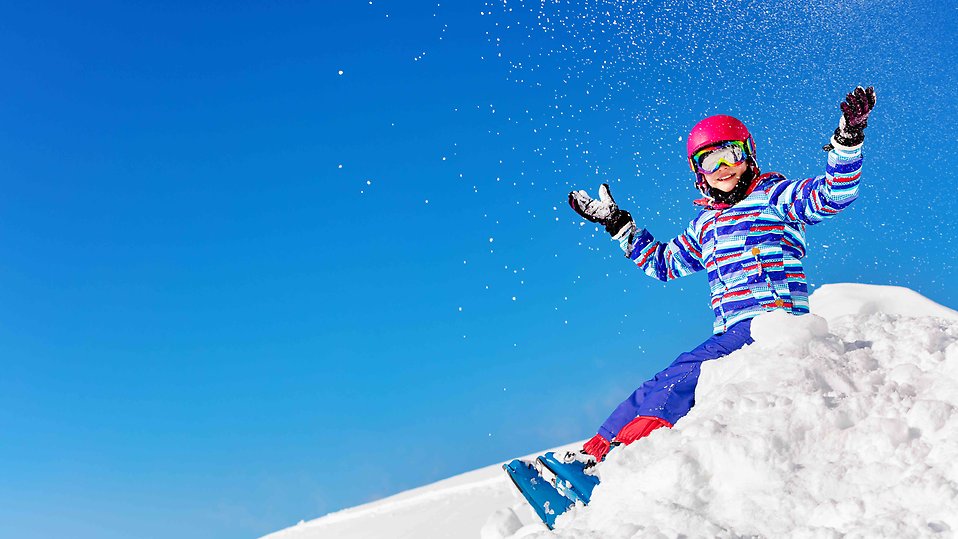 Flicka i skidpjäxor och skidglasögon sitter i snöhög och kastar upp snö. Foto. 