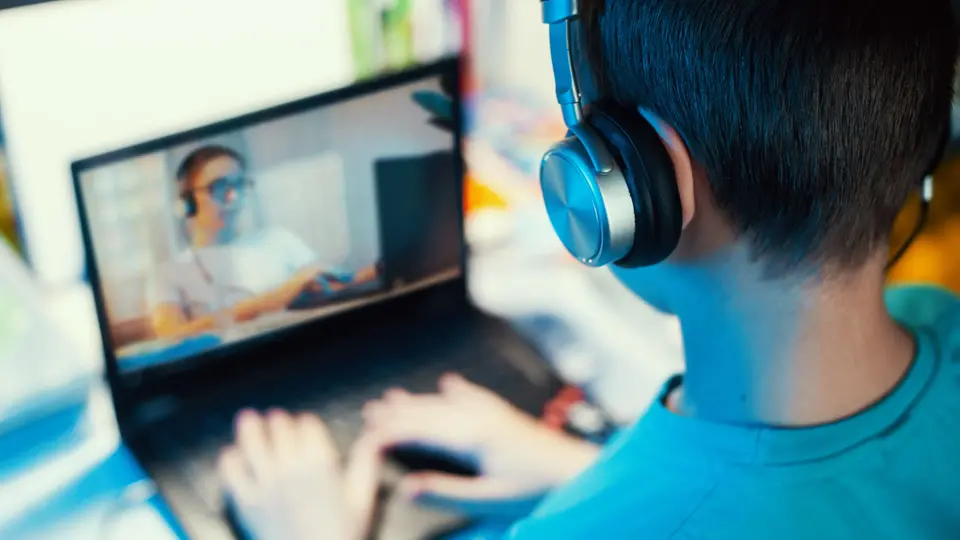 Pojke sitter framför dator med headset. Lärare syns på dataskärmen. Foto. 